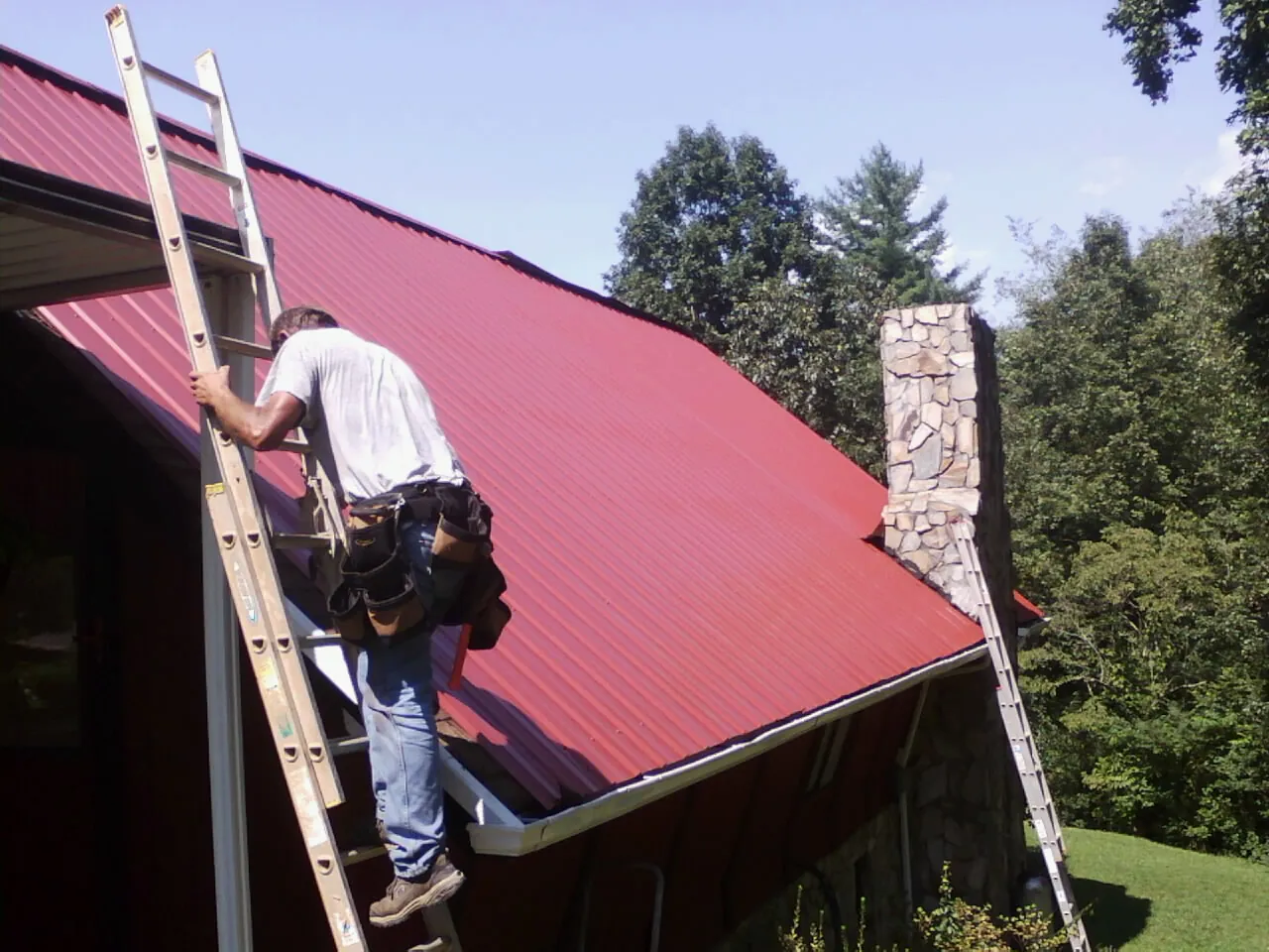 Roofer on a ladder beside a red metal roof and stone chimney — Soffit & Fascia Repair by Roofing Brothers in Granada Hills